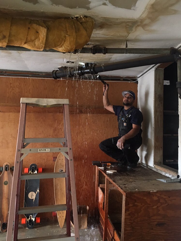 Professional plumber with a flashlight inspecting a major water leak from a ceiling pipe in a residential basement.